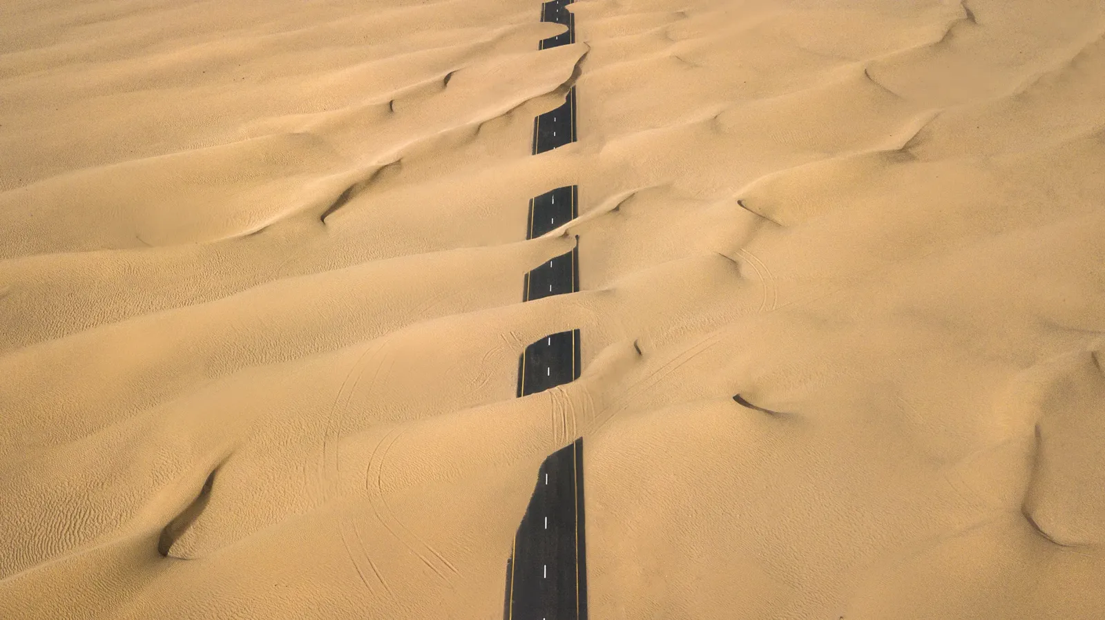 A paved road disappearing under wind-blown sand dunes, seen from above.