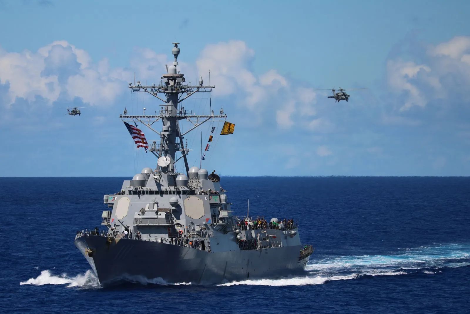 A naval destroyer underway at sea, helicopters overhead.