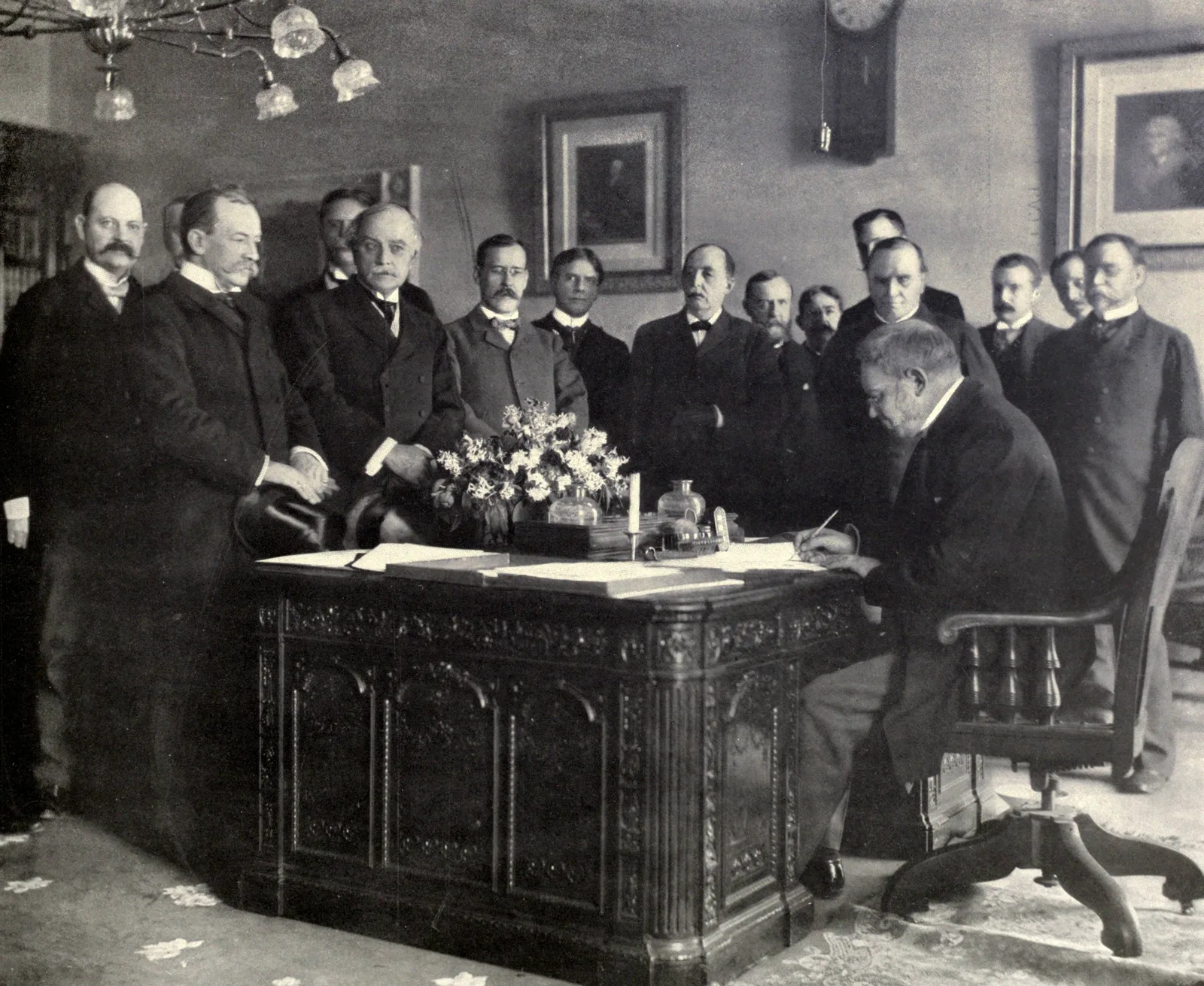 Photograph by Frances Benjamin Johnston of Jules Cambon, the French ambassador to Washington who negotiated on Spain's behalf, signing the memorandum of ratification of the peace settlement in 1899, watched by American officials. The Treaty of Paris itself was signed in Paris on 10 December 1898; this photograph shows the ratification step that followed it.