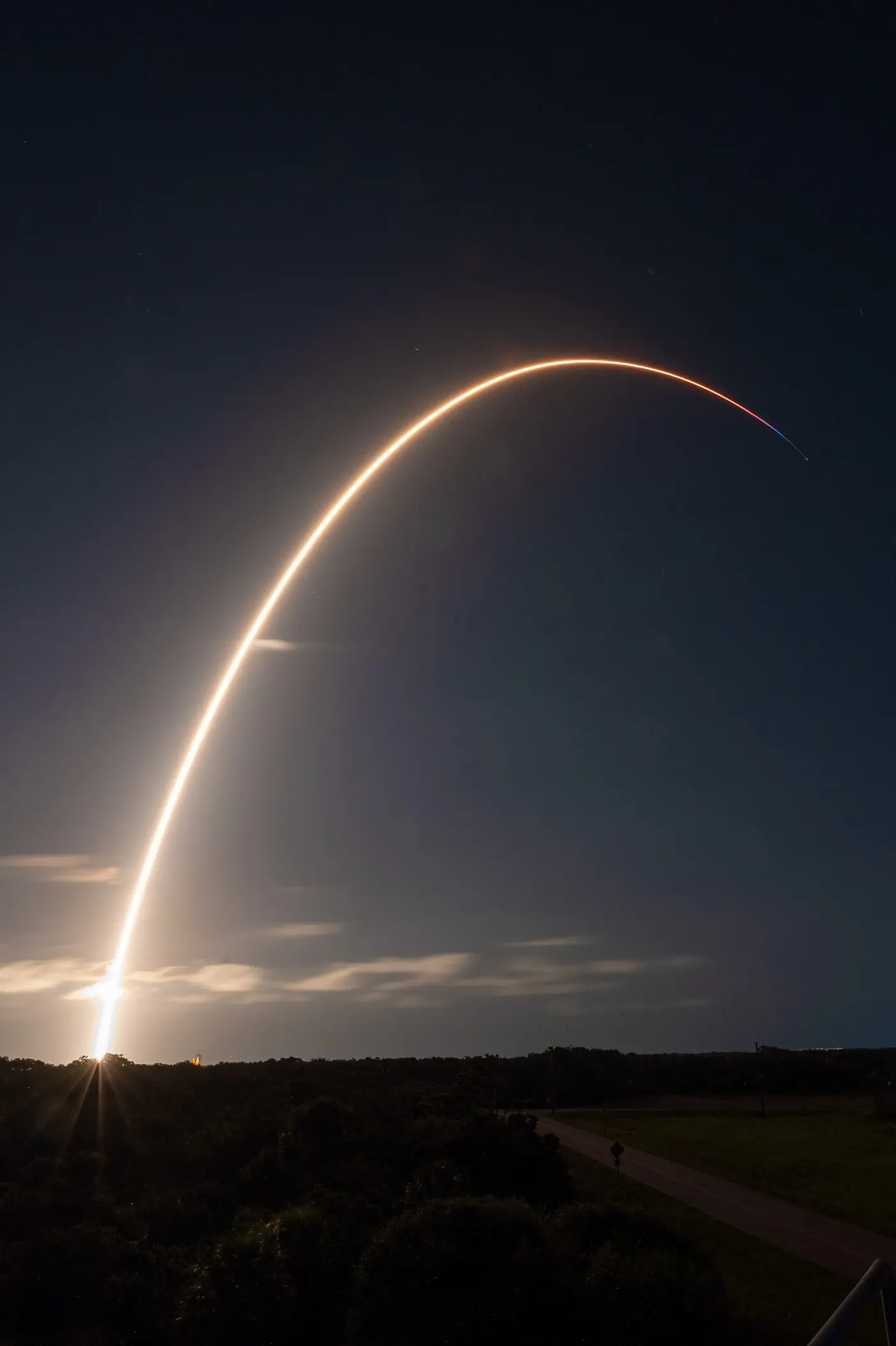 A long-exposure photograph of a Starlink satellite launch at night, its exhaust trail arcing over dark ground (file photo). This photograph does not depict any event reported in this article.