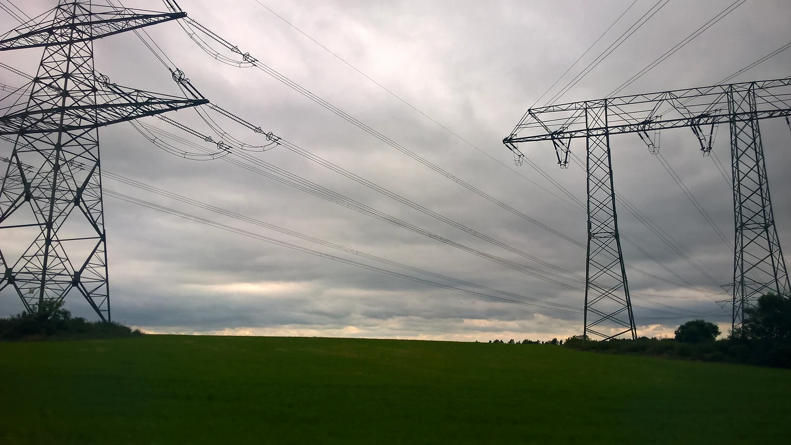 High-voltage electricity pylons and power lines crossing a green field under a cloudy sky. Generic photo; it does not depict the events described in this article. — Image: Lupus in Saxonia / Wikimedia Commons (CC BY-SA 4.0)