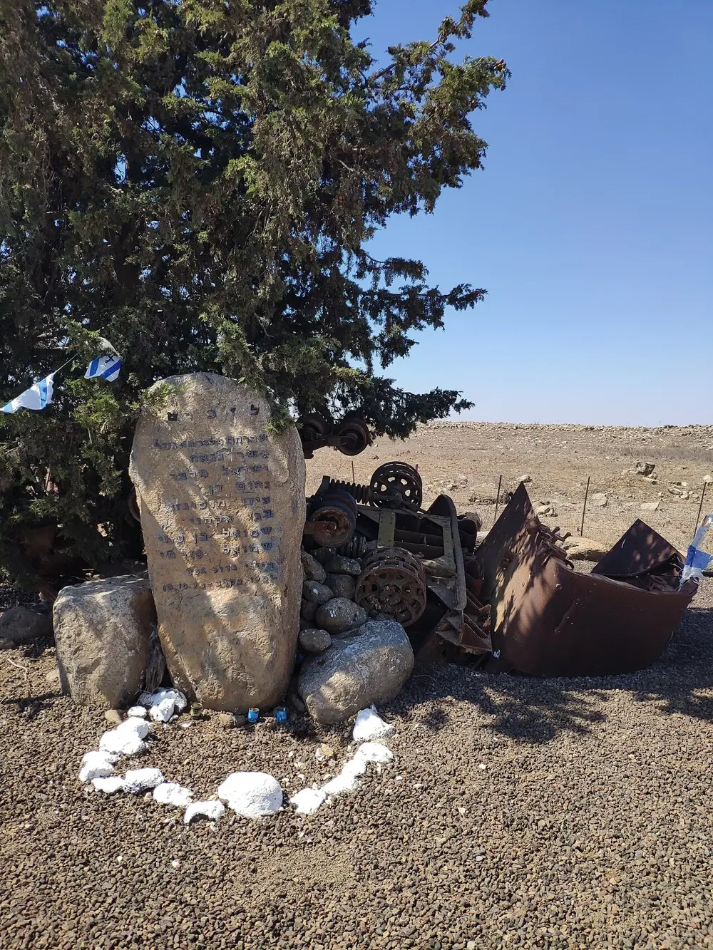 A battalion memorial on the Golan Heights, photographed in modern times: an inscribed stone, flags, and preserved wreckage kept as part of the memorial. The Golan was attacked by Syria on 6 October 1973. This is a modern memorial photograph; it does not depict the fighting.